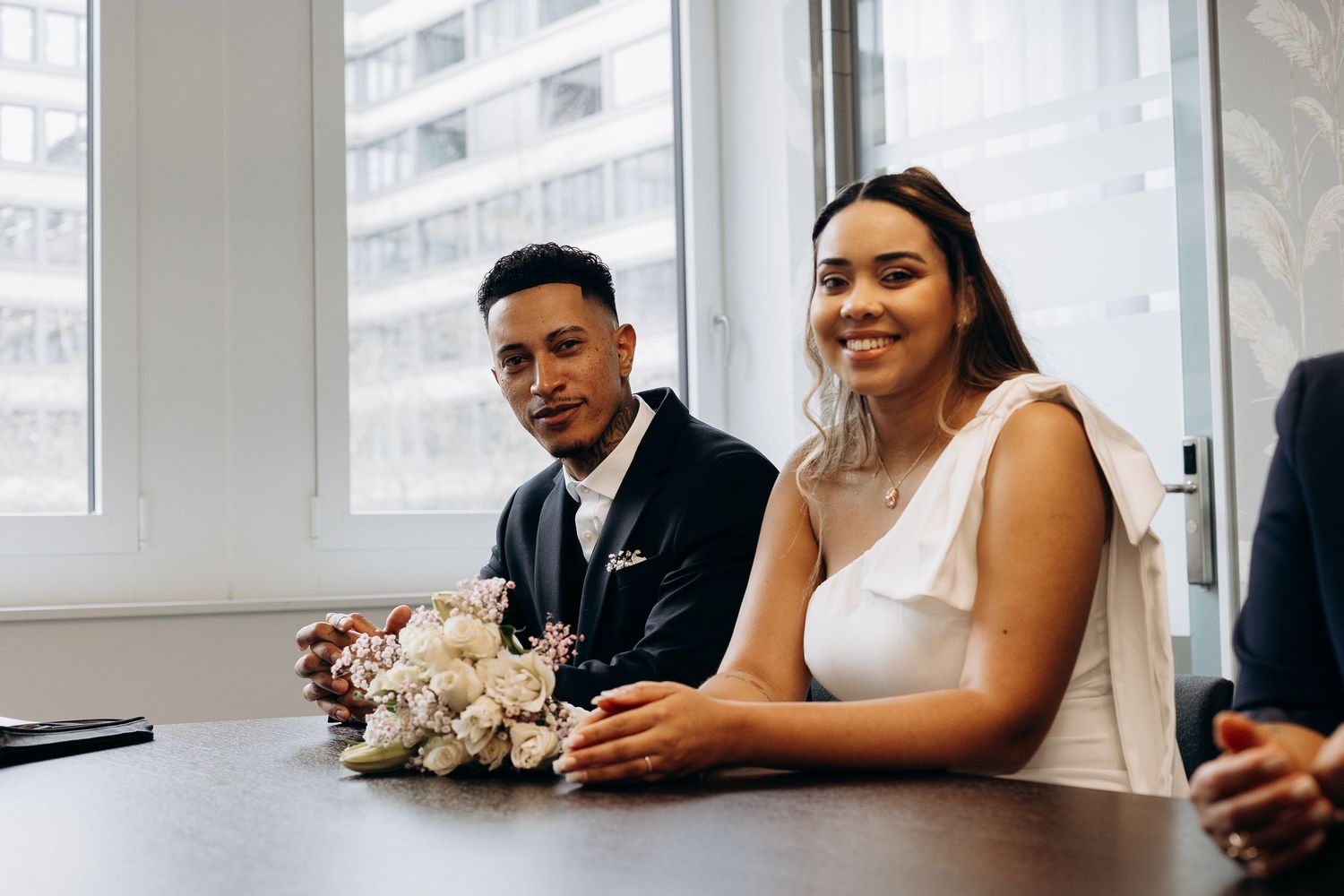 A couple sits at a table during their civil marriage ceremony with a white floral bouquet.