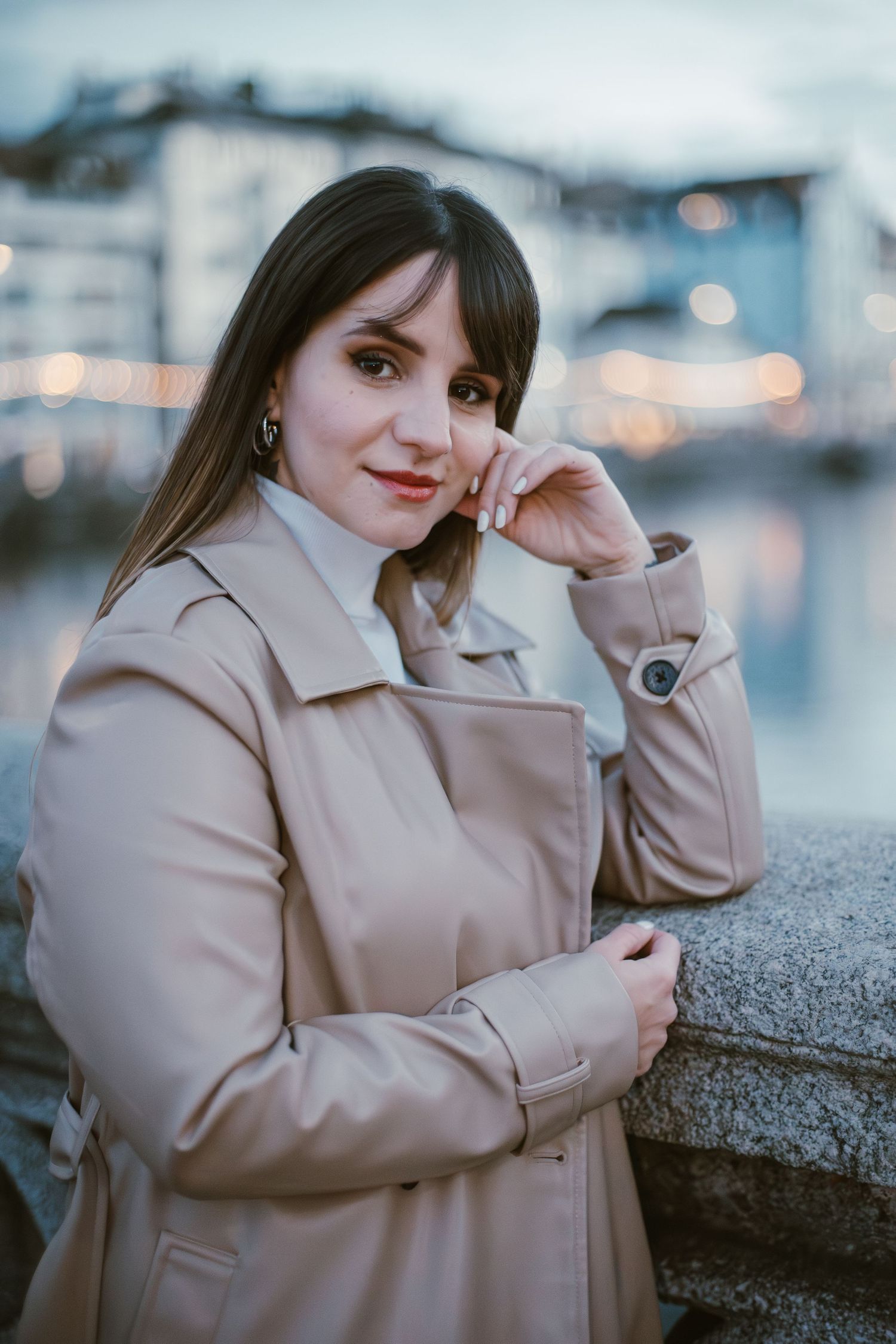 Woman in beige coat poses against urban waterfront setting during blue hour.