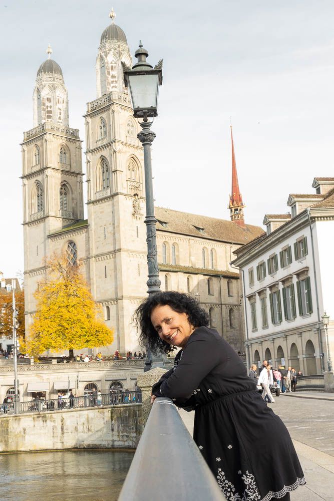 Person leaning on railing with Grossmünster cathedral and autumn trees in background.