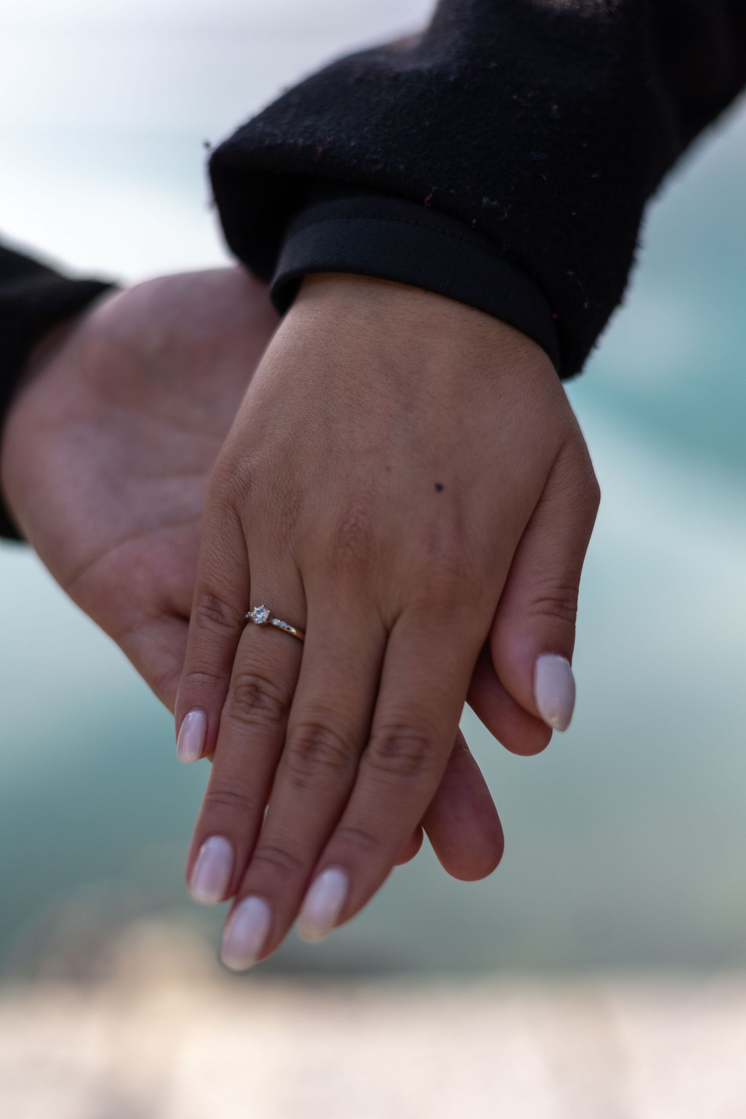 A close-up shot of hands showing off a diamond engagement ring against a blurred waterfront background.
