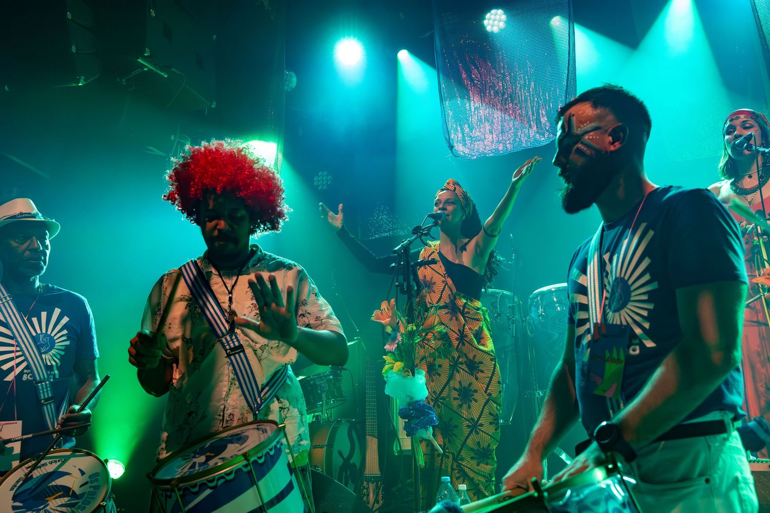 Musicians in colorful costumes perform on stage under vibrant blue-green lighting.