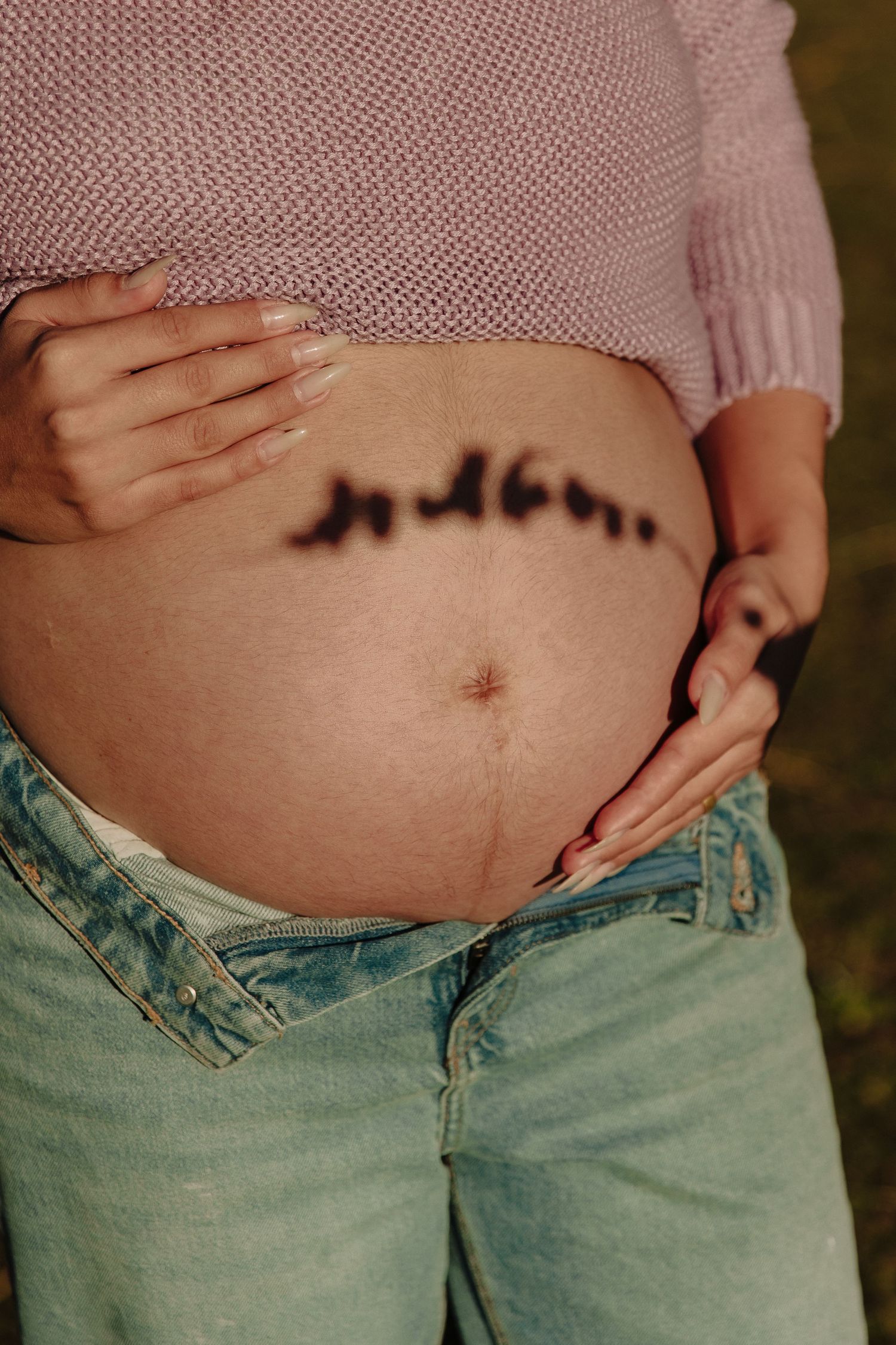 Close-up of pregnant belly with text written across it, wearing pink sweater and mint jeans.