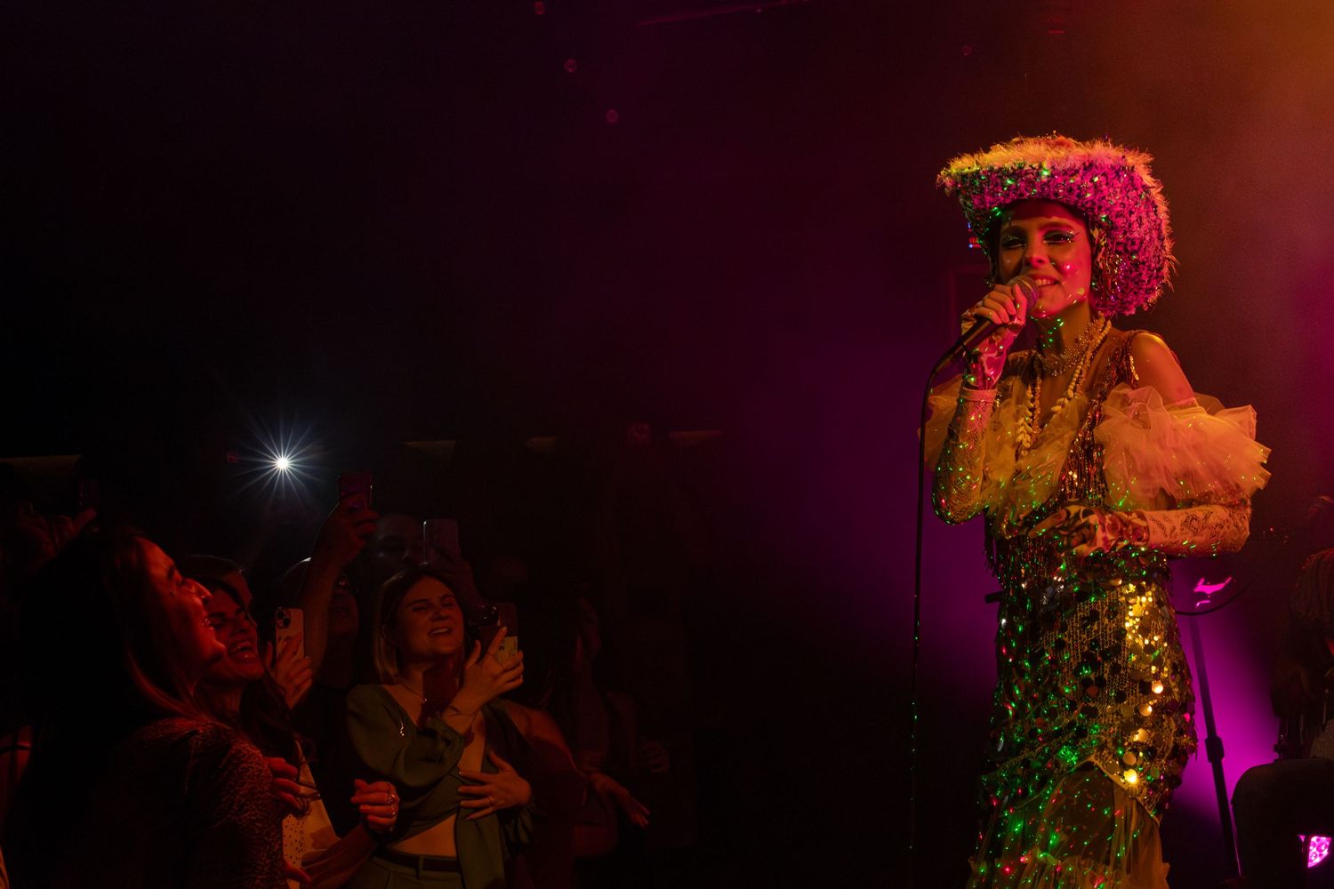 A glittering performer on stage entertains an energetic crowd under colored lights.