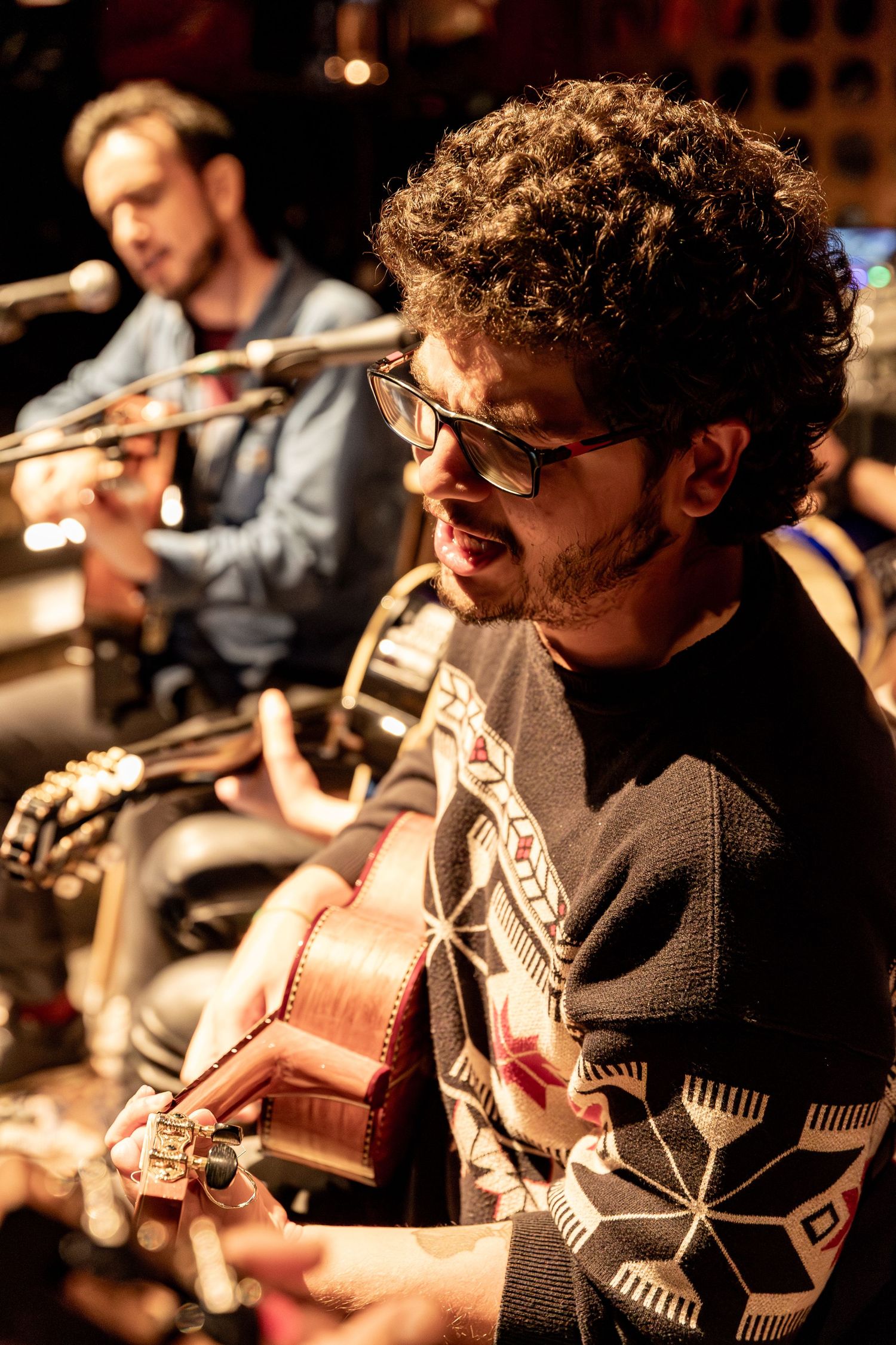 Musician playing acoustic guitar during intimate performance wearing patterned sweater.
