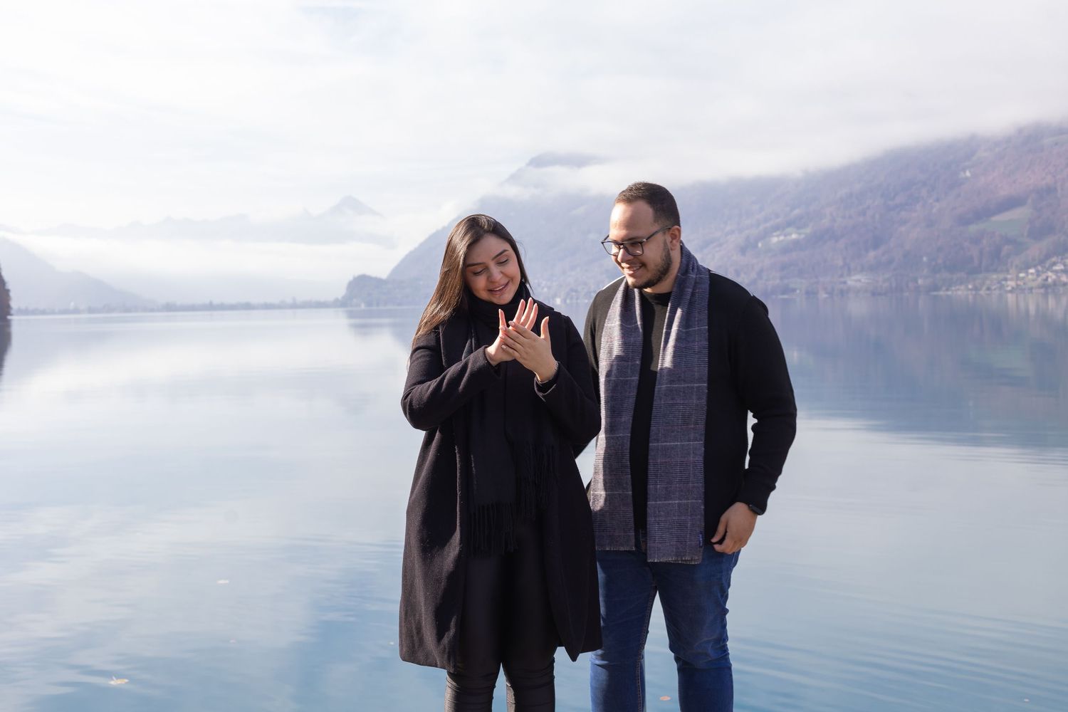 Newly engaged couple shares excited reactions on dock with scenic mountain lake view.