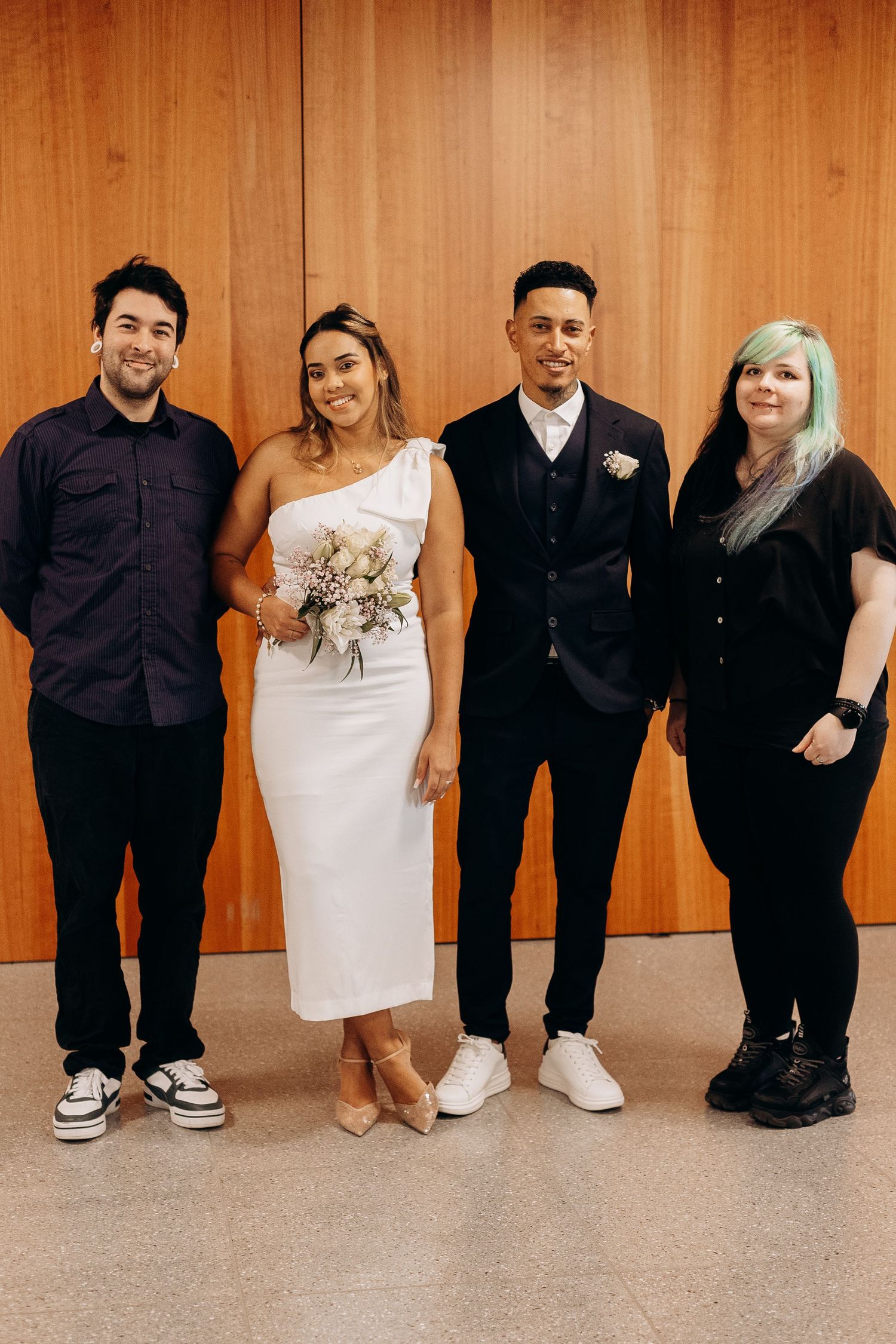 Small wedding party poses against wood paneled wall in modern formal attire.