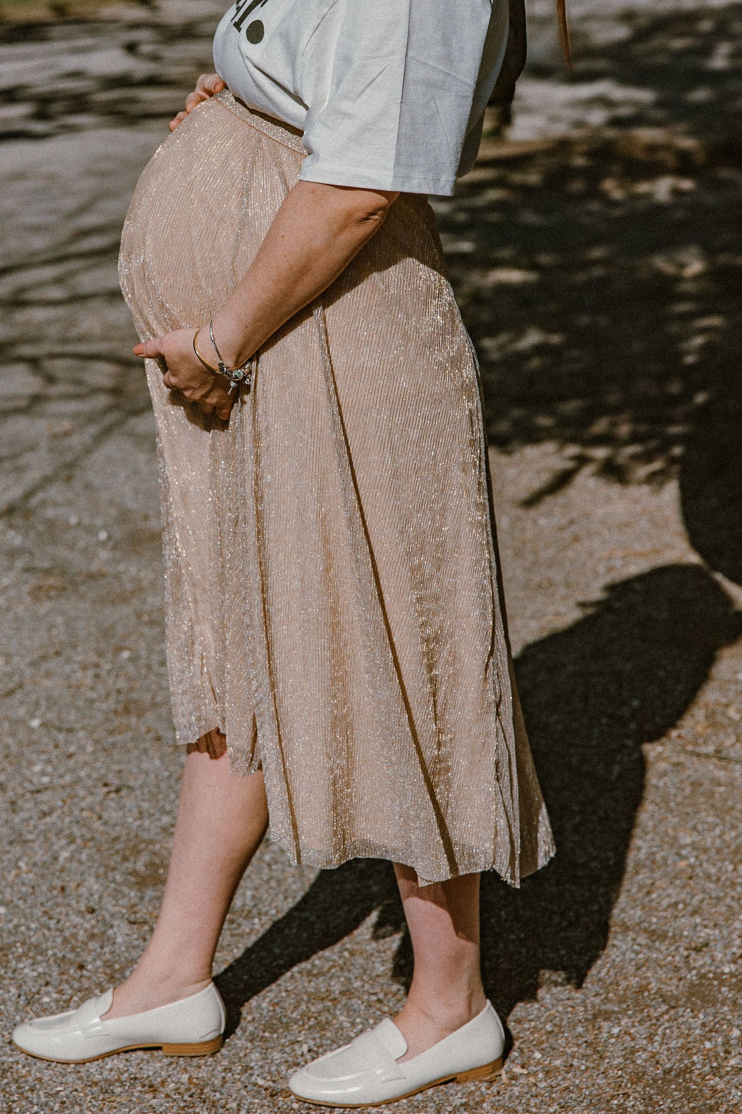 Side view of pregnant woman in flowing beige skirt and white flats standing in dappled sunlight.