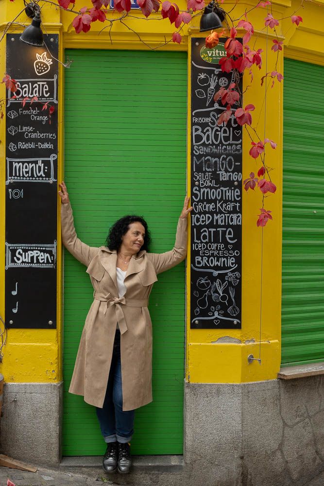 Person in tan coat poses playfully against vibrant yellow and green storefront with menu boards.