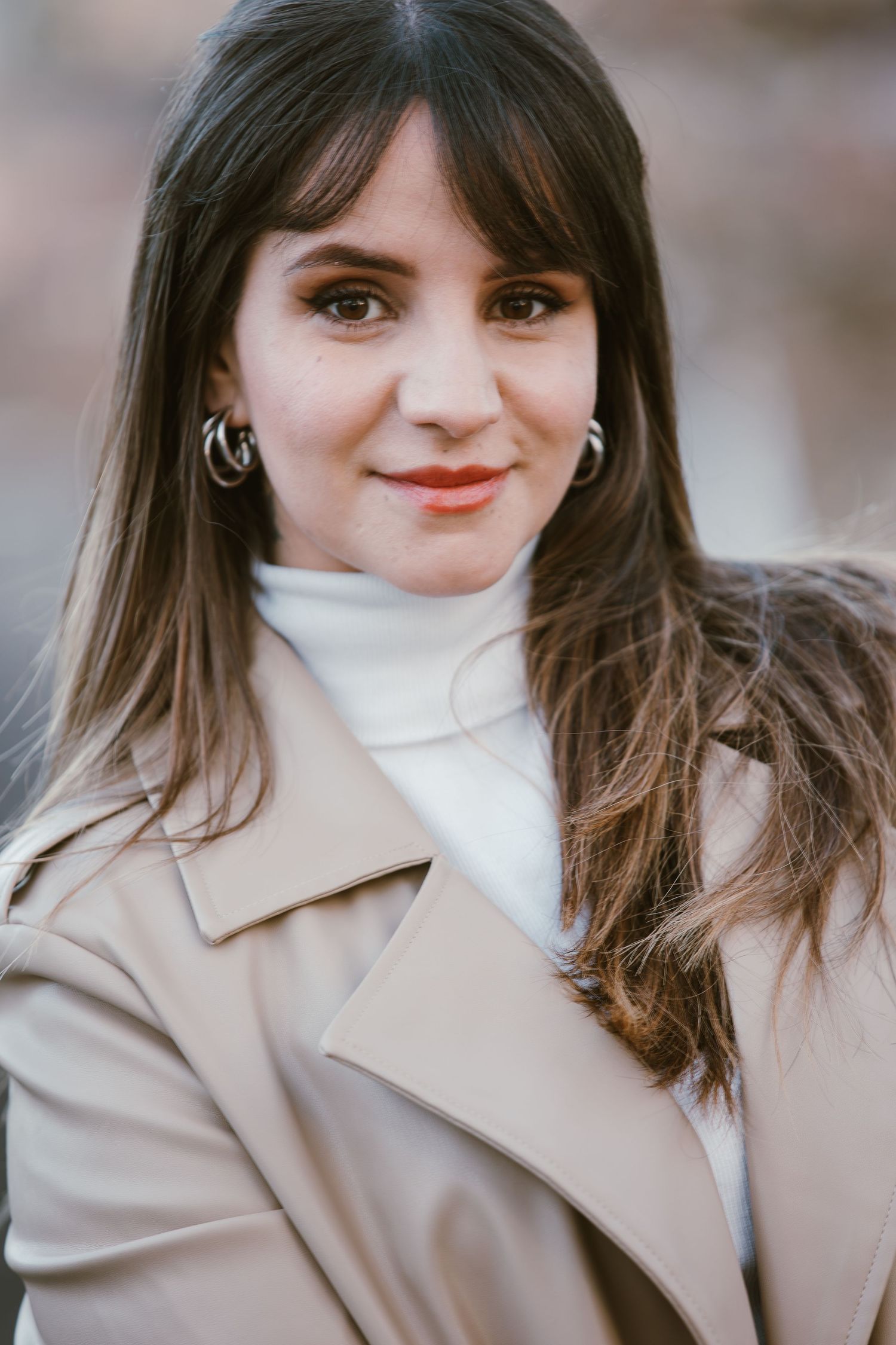 Portrait of a woman in a beige coat and white turtleneck with long dark hair.