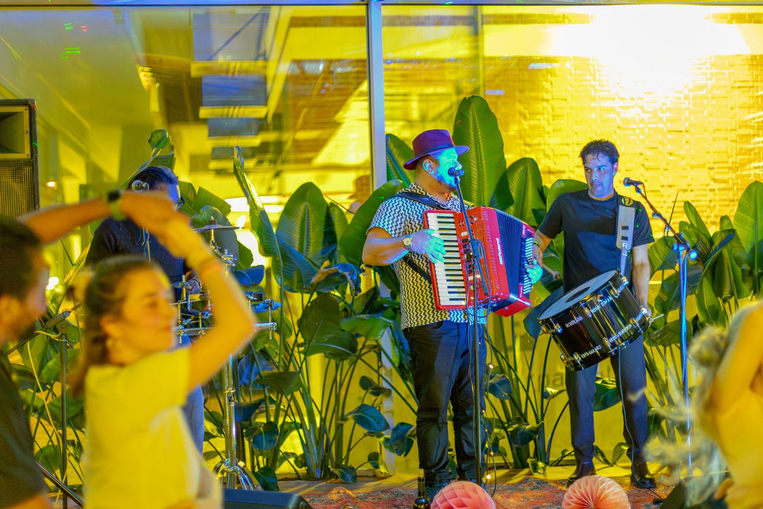 Musicians perform on stage with accordion among yellow lights and tropical decor.