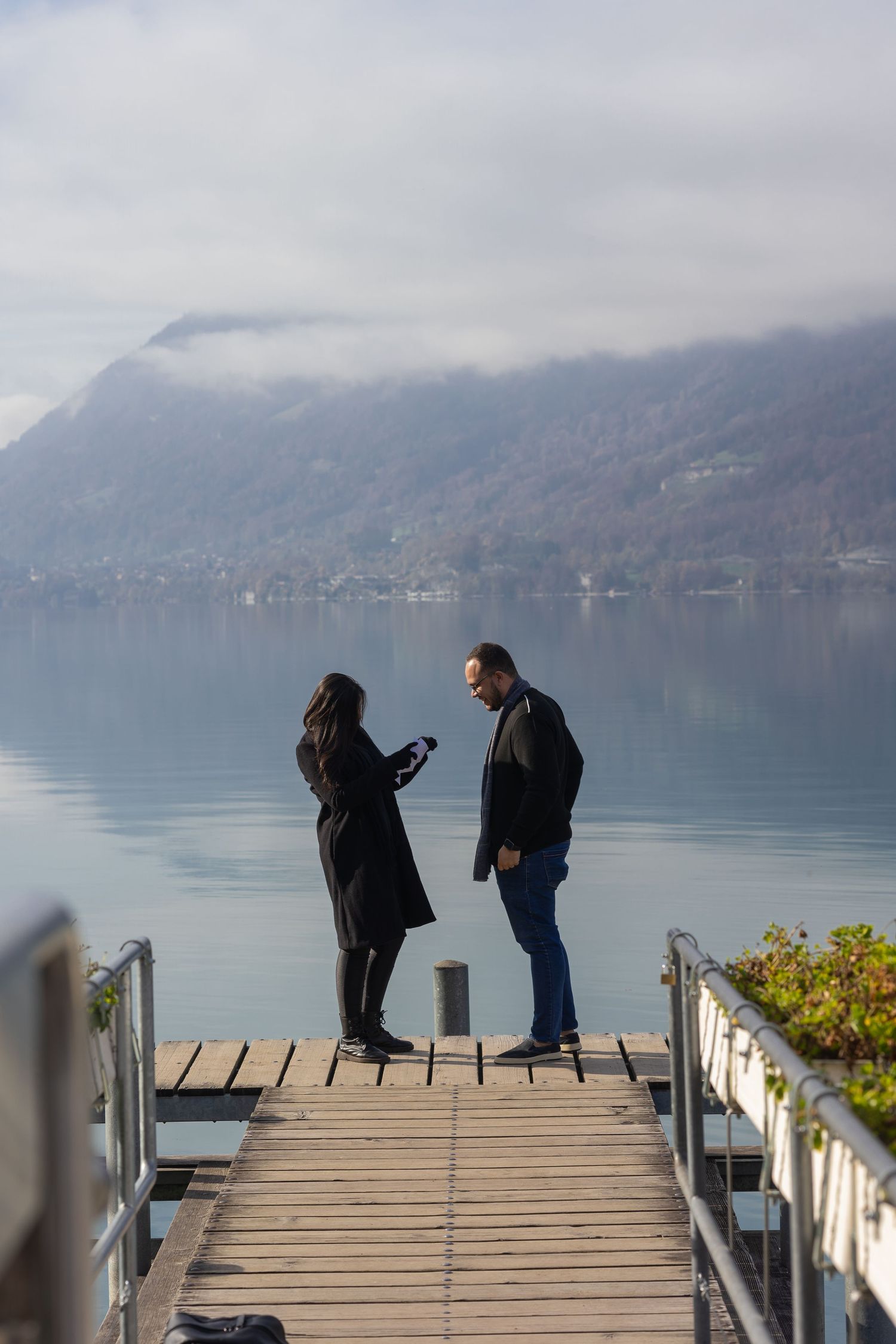 Two people stand on a wooden dock overlooking a misty mountain lake on a calm winter morning.