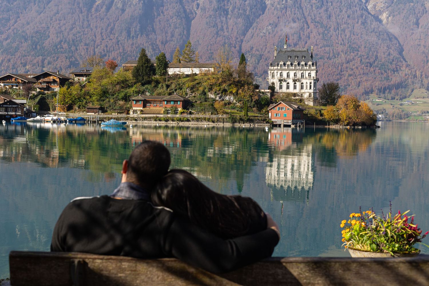 A couple sits on a bench overlooking a grand chateau on a lake with mountain backdrop.