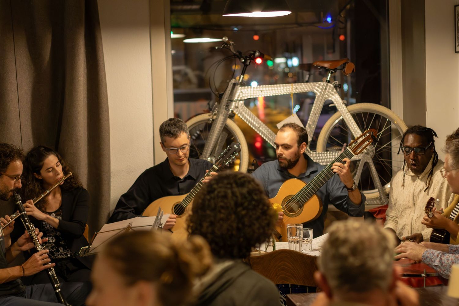 Musicians gather around a table with instruments while a bicycle hangs on the wall in the background.
