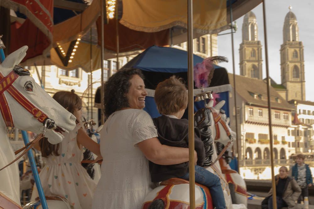 Family enjoying a ride on a vintage carousel at a fair with historic church spires in the background.
