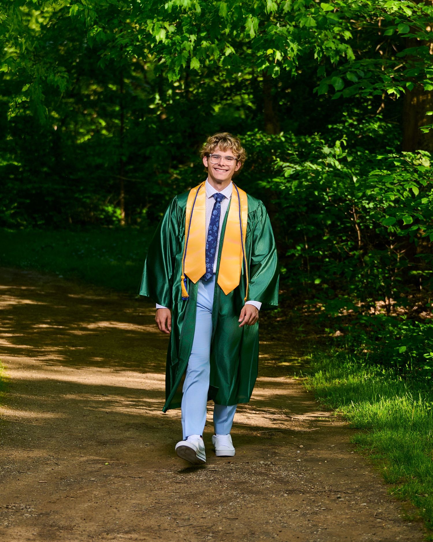 Graduate in green cap and gown standing on dirt path in lush forest setting wearing blue tie and jeans.