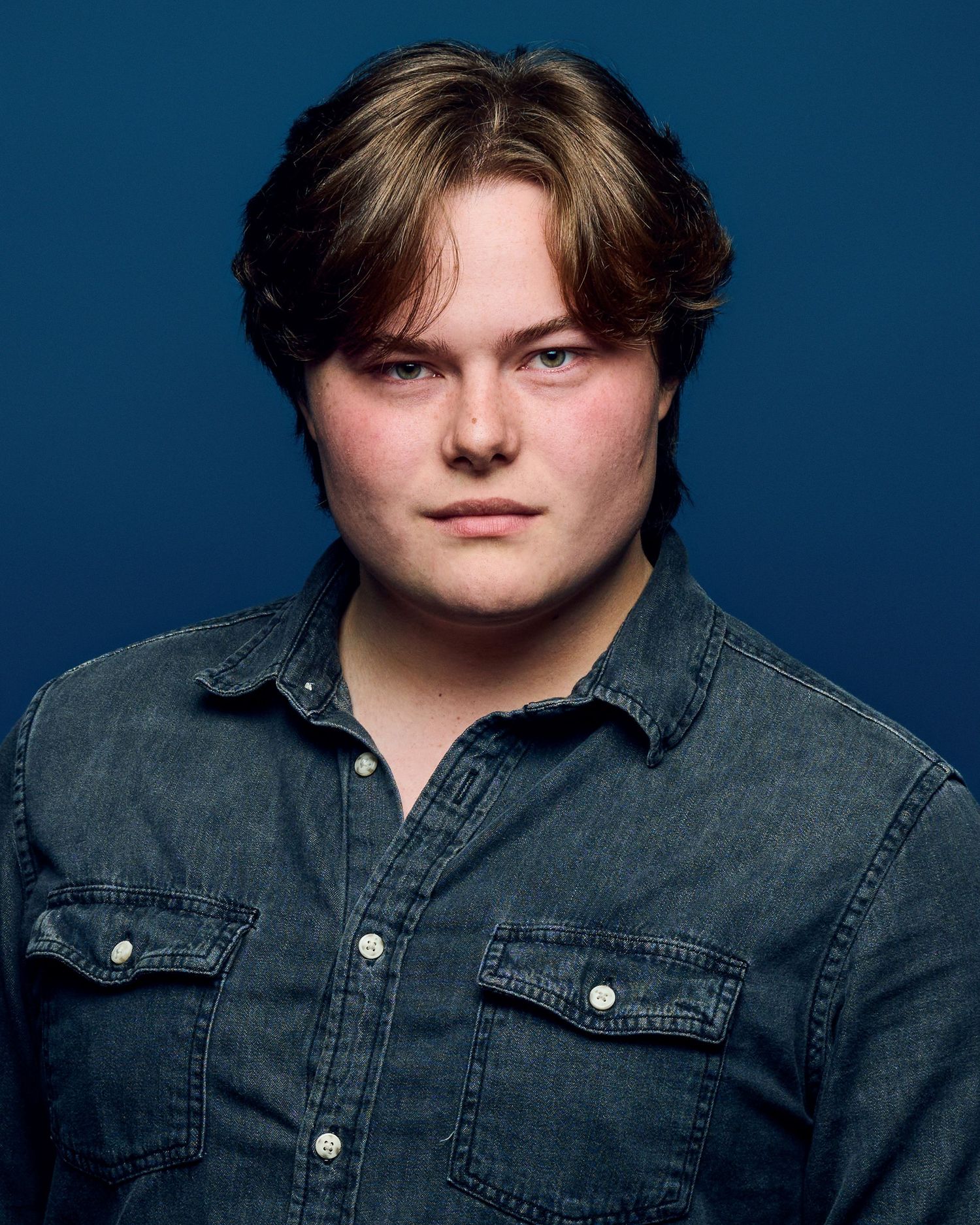 Portrait in denim shirt with layered hair against blue background.