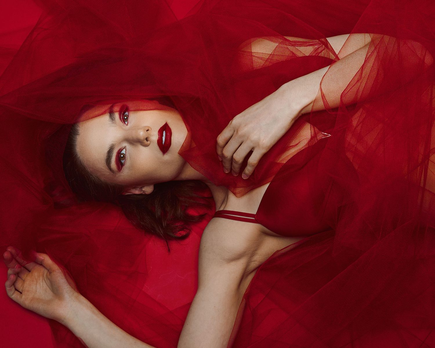 Fashion model in red dress poses dramatically against vibrant red backdrop in artistic portrait photography.