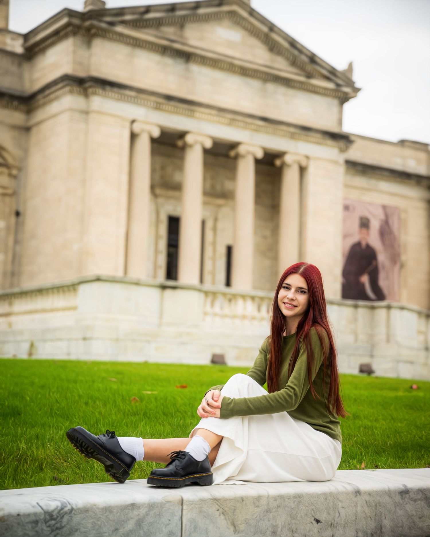 Student sitting casually on steps of a neoclassical building wearing a green sweater and white skirt.