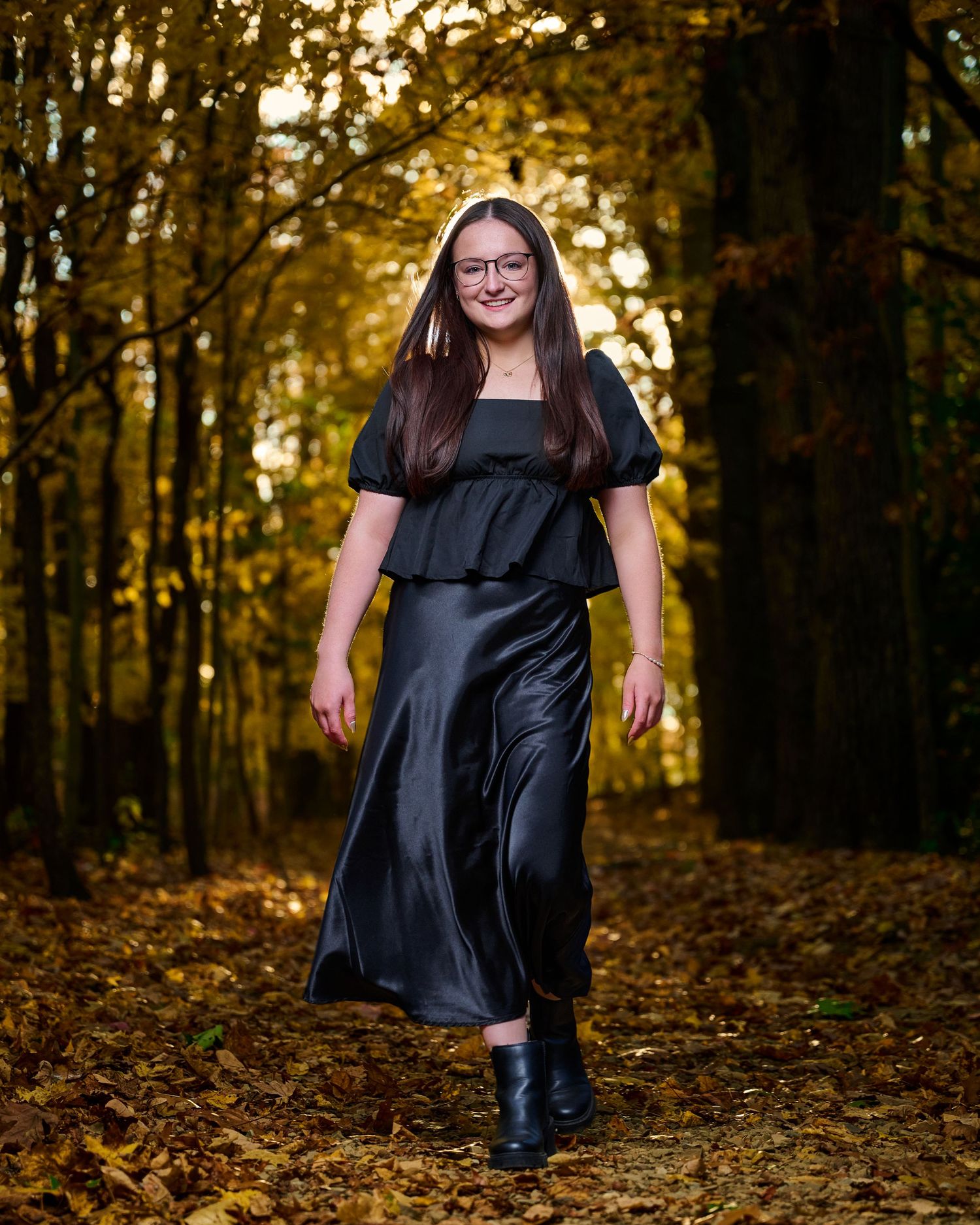A fashion portrait of someone in a black dress walking through autumn woods with golden sunlight filtering through trees.