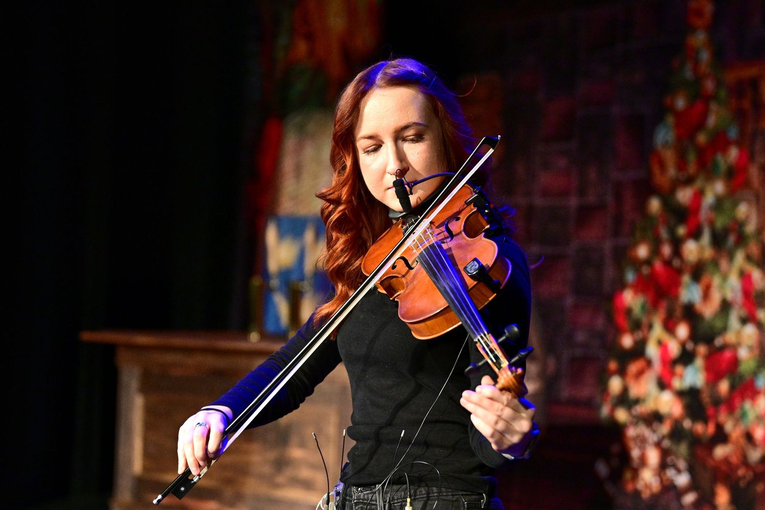 Violinist in black attire performs passionately against a festive backdrop with twinkling lights.