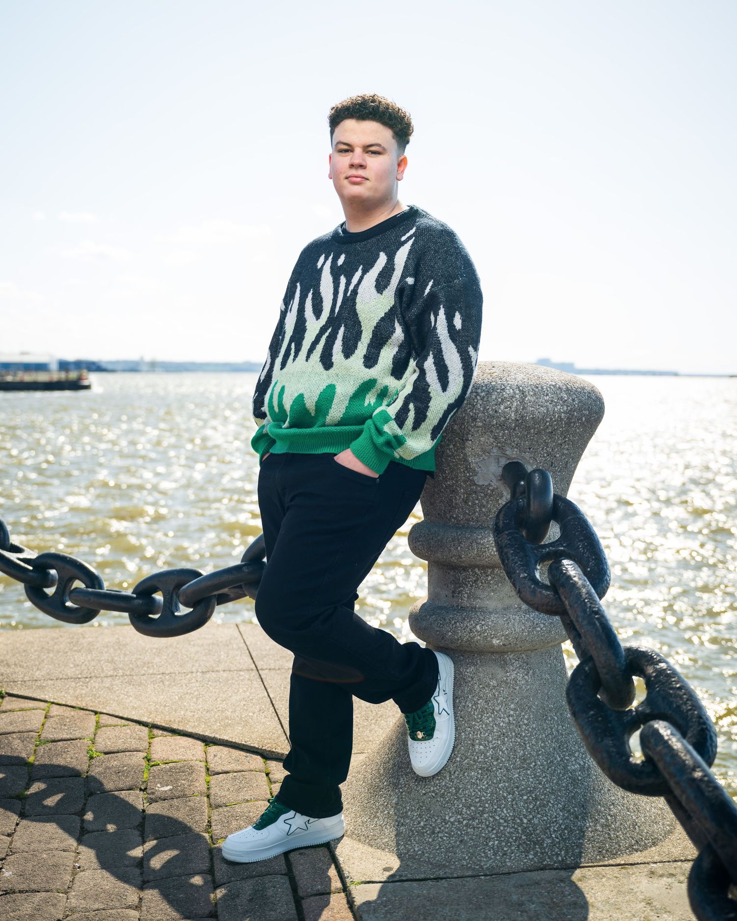 An urban portrait of someone in a flame-print sweater posed by waterfront chains and concrete bollards.