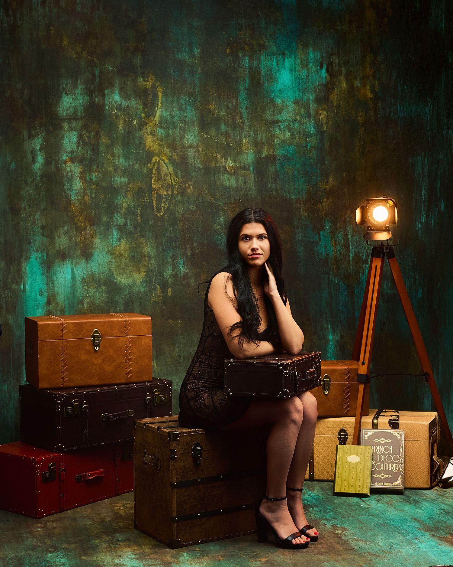 Artistic portrait against teal textured wall with vintage suitcases and lantern creating moody atmosphere.