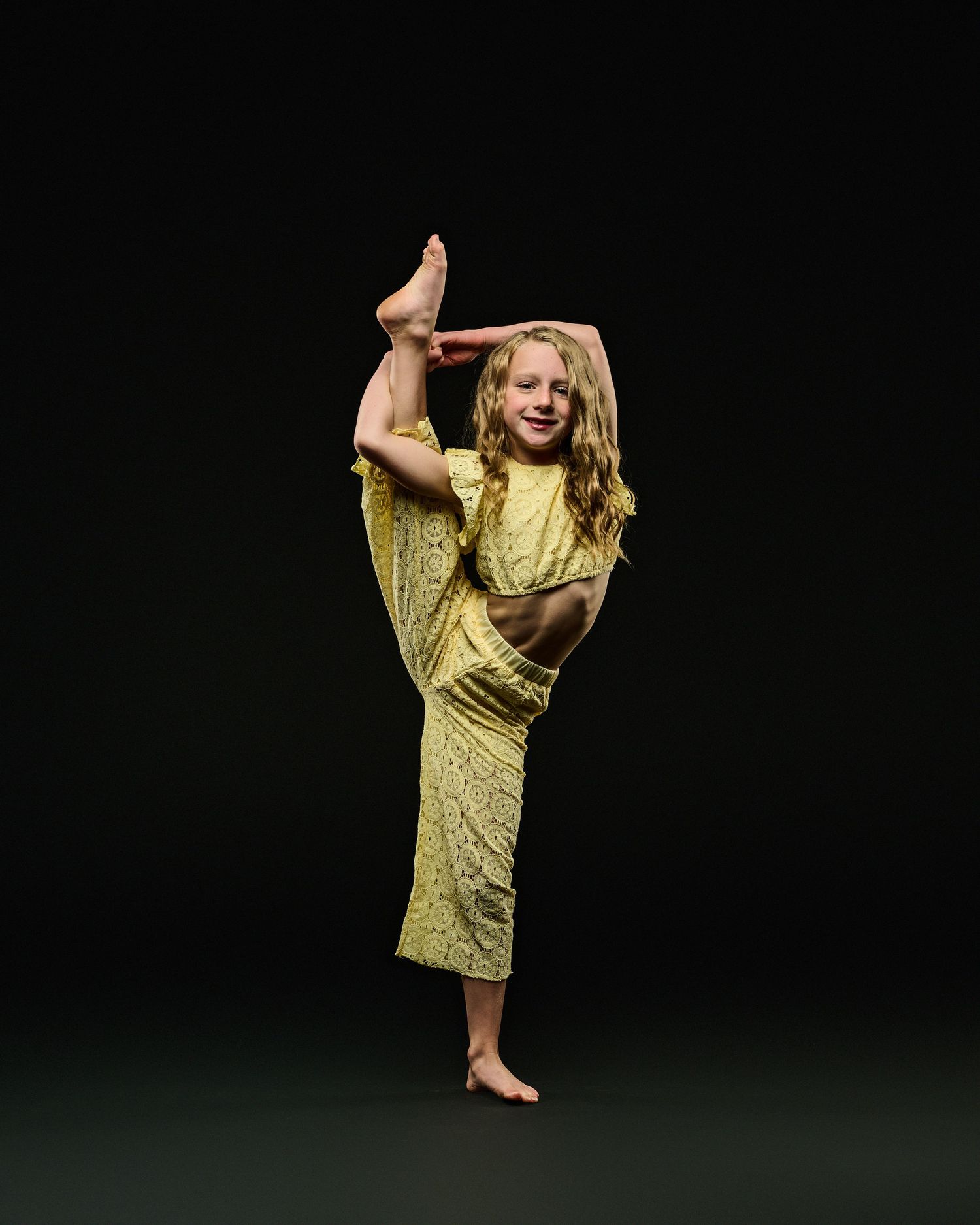 A dancer in yellow performs a high leg extension against a dark studio background.