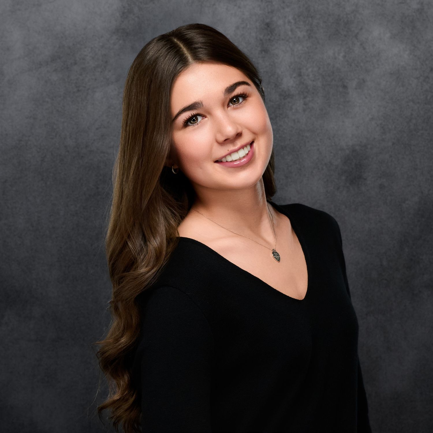Professional headshot with long brown hair wearing black v-neck top against grey background.