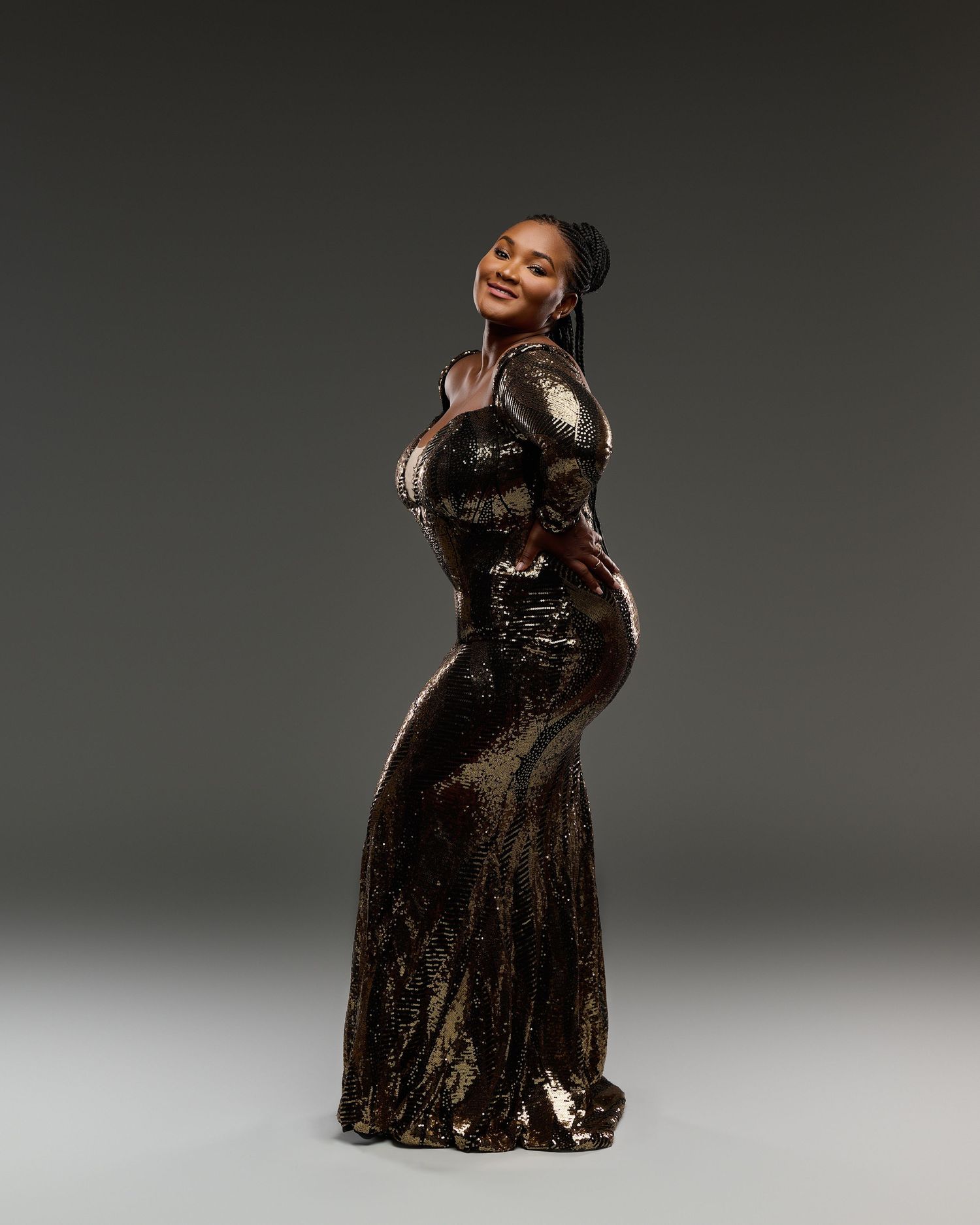 Figure in shimmering gold evening gown poses confidently against dark background.