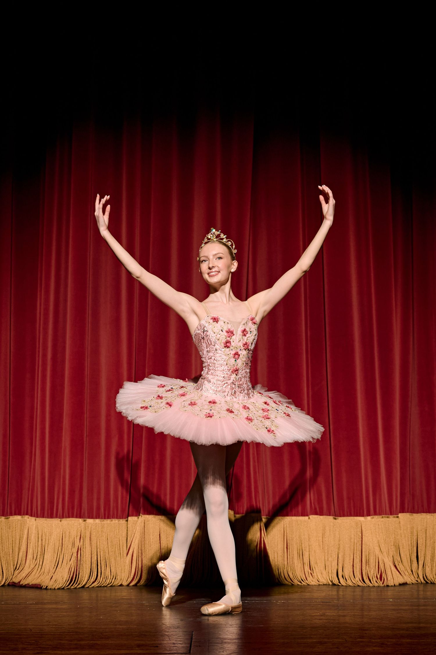 A ballerina poses in a pink tutu against red curtains on a theater stage.