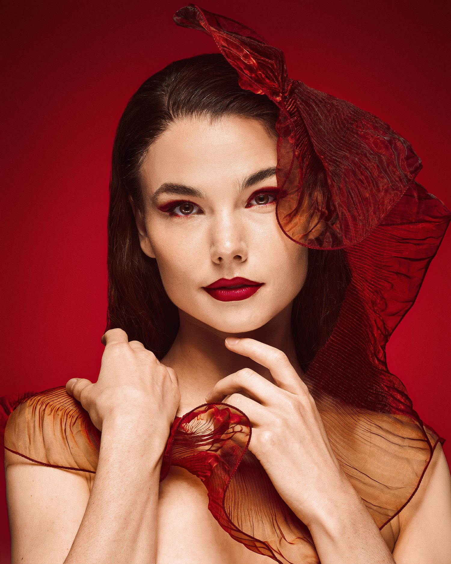 Beauty portrait with flowing red fabric and dramatic makeup against crimson backdrop.