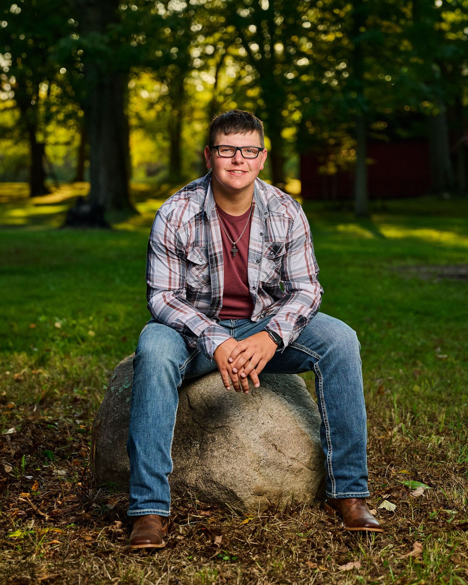 A relaxed outdoor portrait of someone sitting on a rock wearing casual plaid and denim in a park setting.