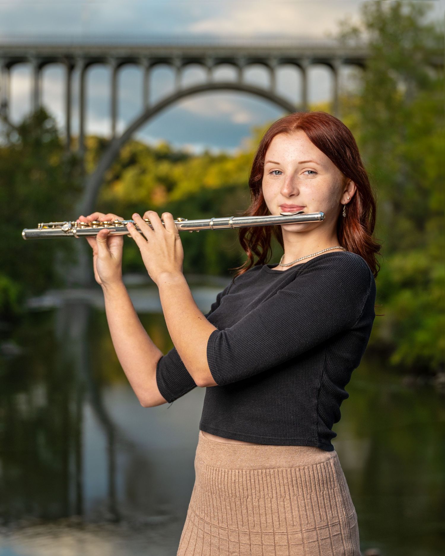Musician playing flute outdoors with arched bridge in background.