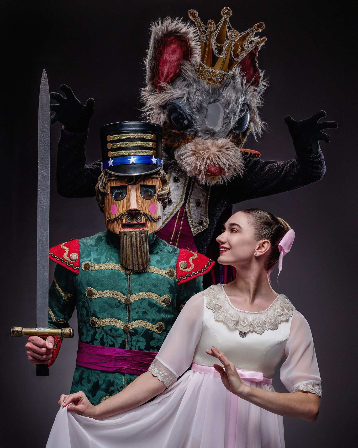 A ballet dancer in white dress poses next to a towering Nutcracker figure with crown and sword against dark background.