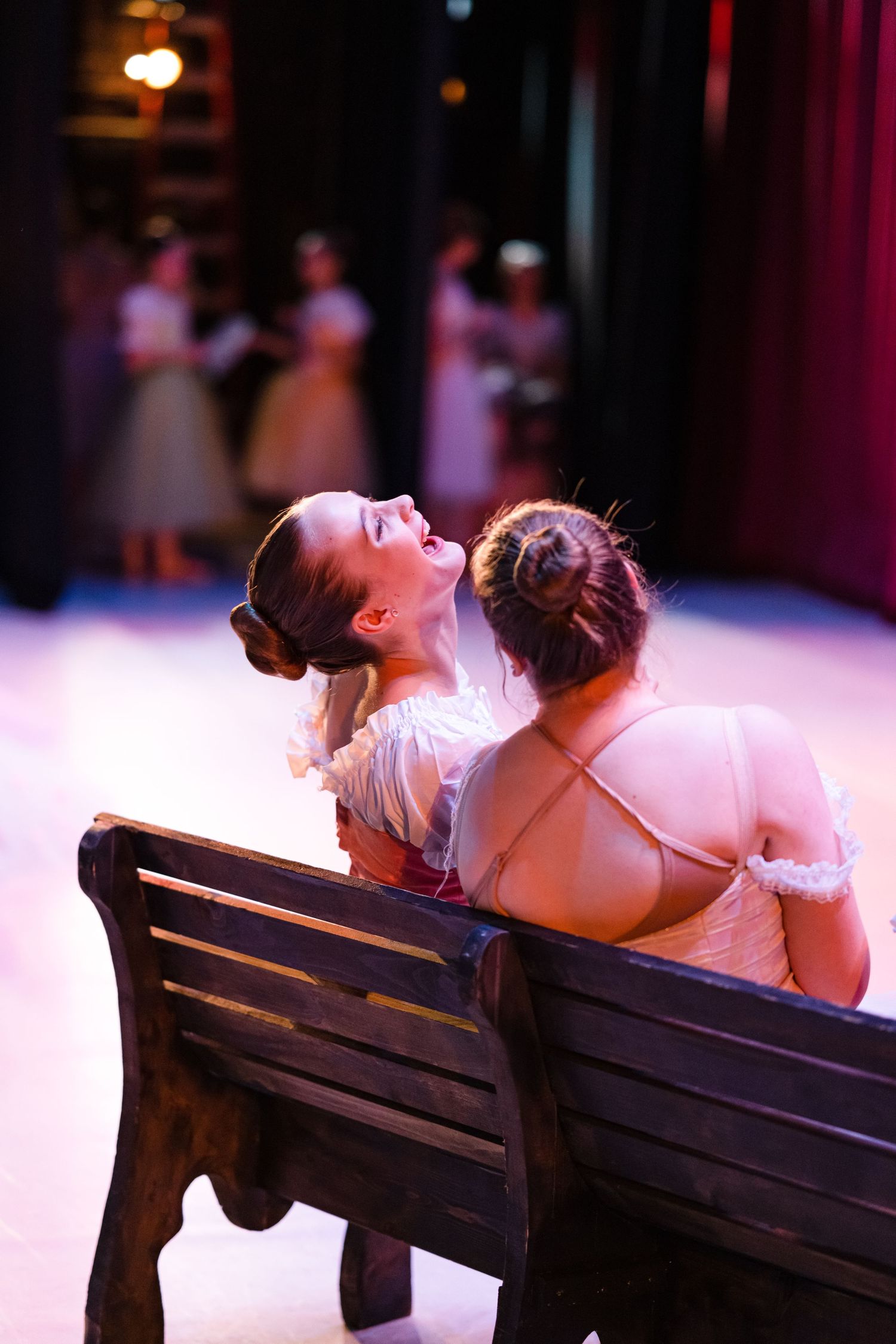Two dancers in white costumes share a moment while sitting on a wooden bench.