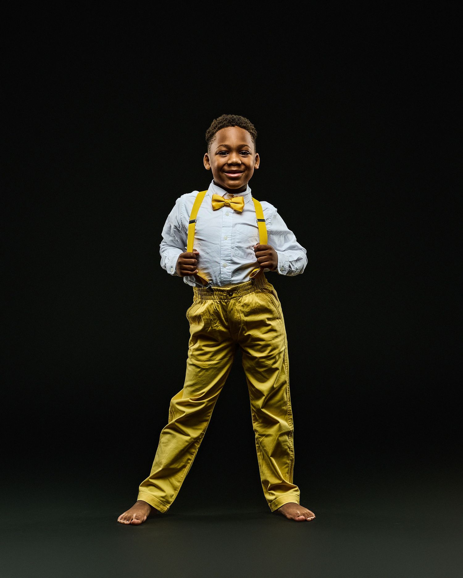 A smiling performer in yellow suspenders and bow tie stands confidently against a black background.