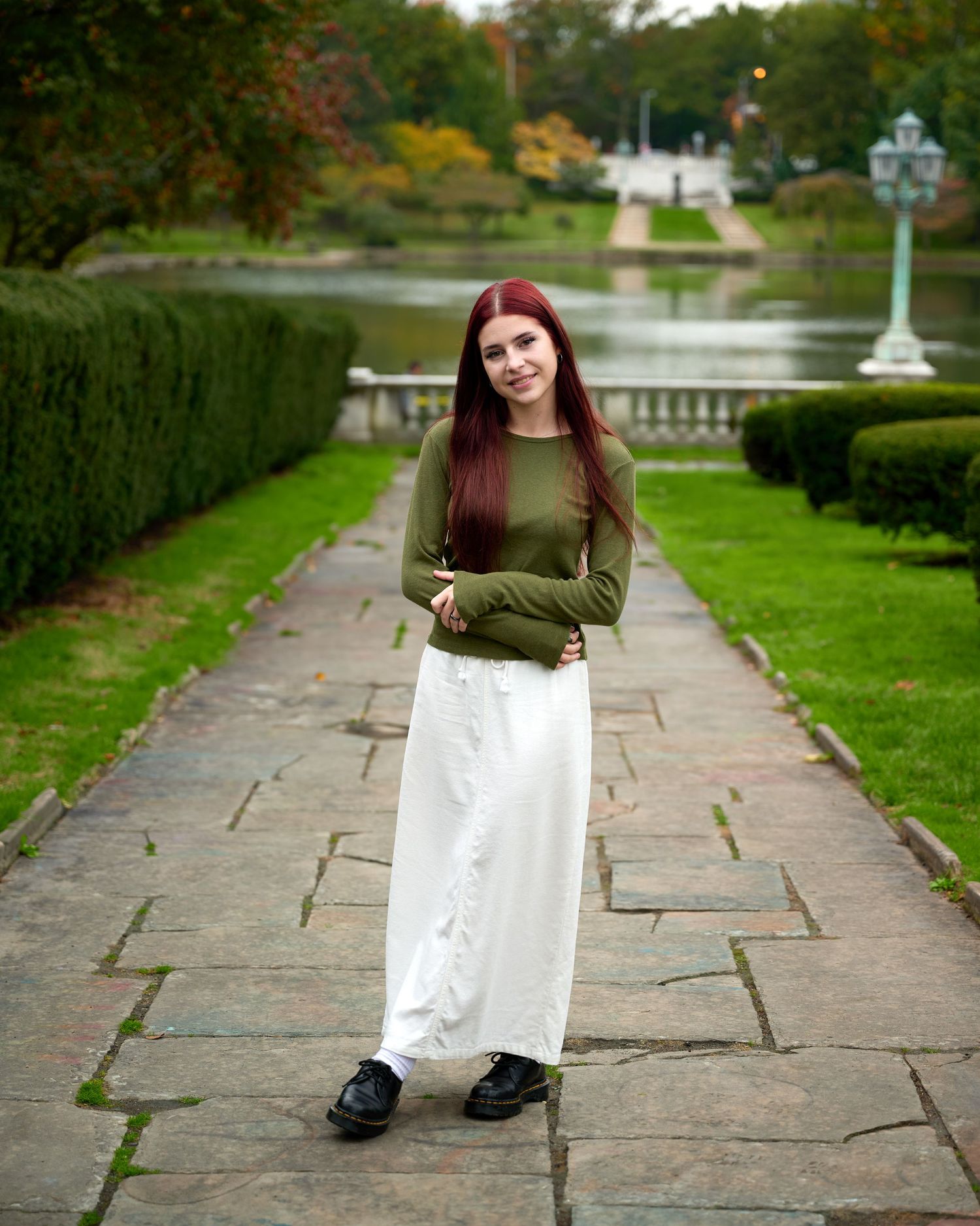 Figure in olive sweater and white pants standing on garden path with pond view.