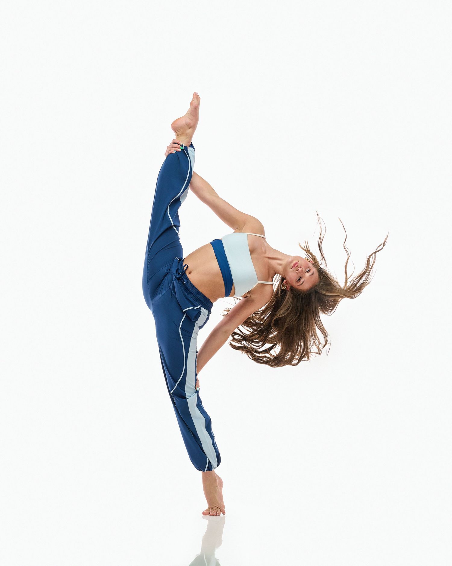 A dancer in blue athletic wear performs a dynamic high kick with flowing hair movement against a white background.