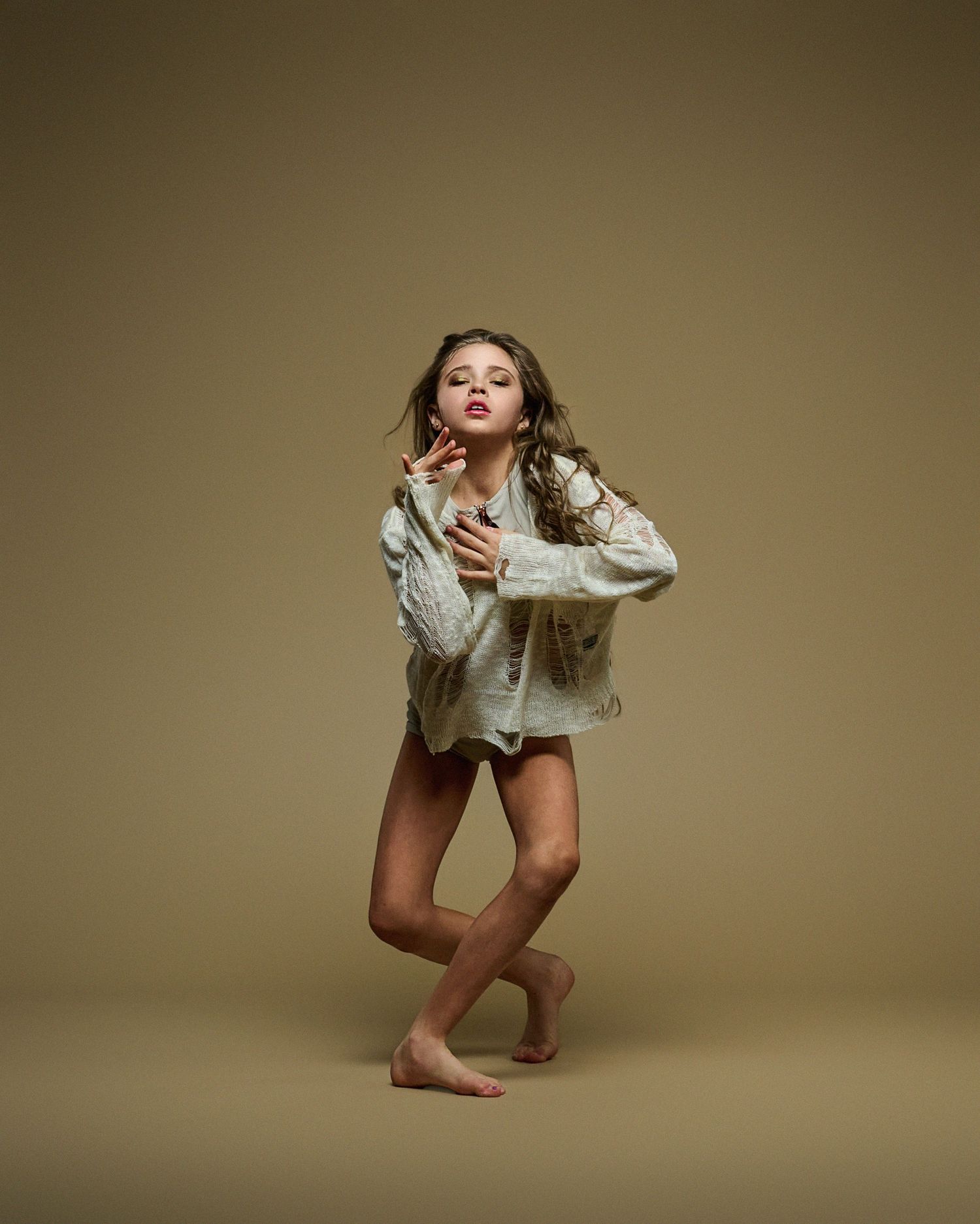 Dance performer in oversized white shirt striking dramatic pose on brown backdrop.