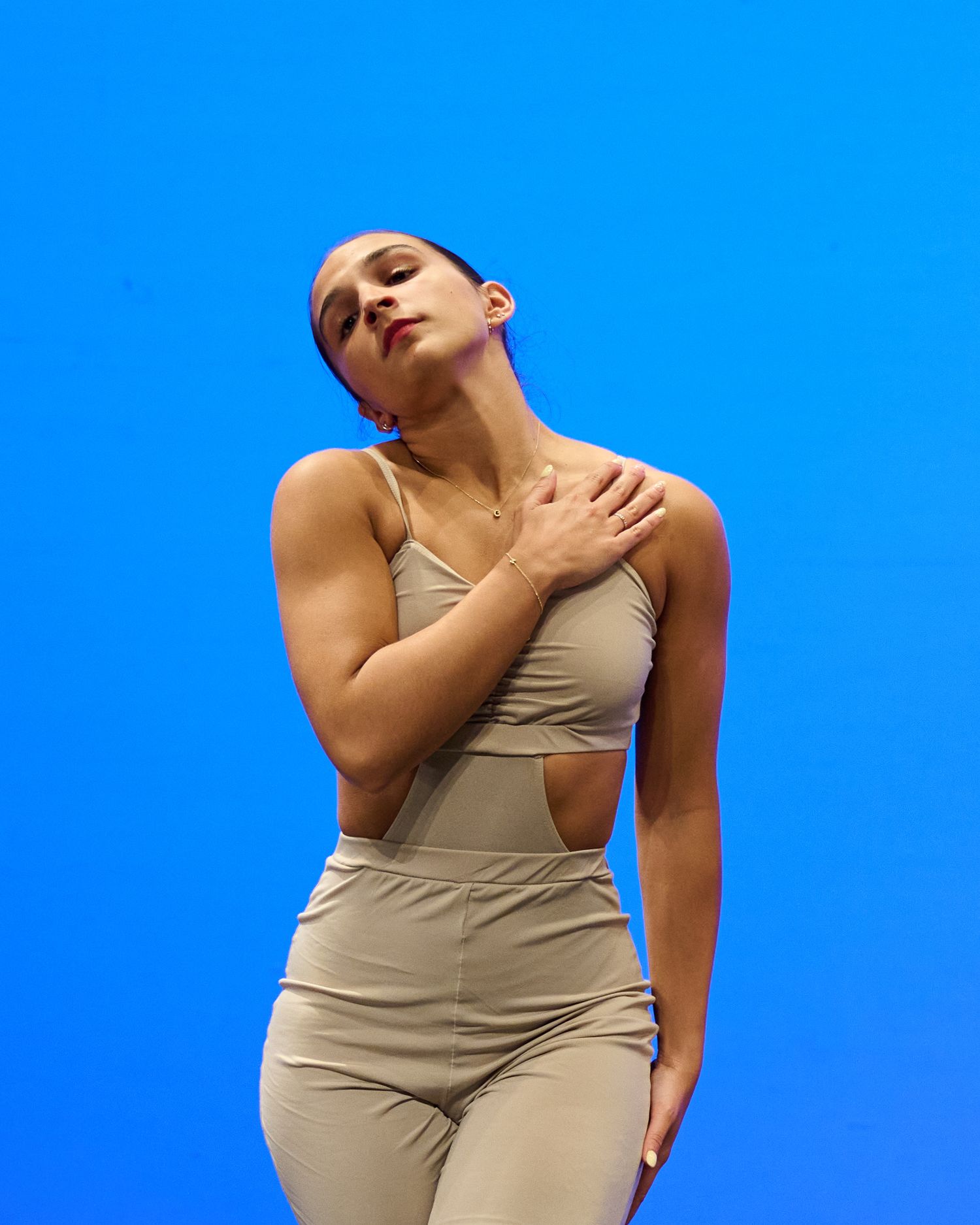 Dancer in beige outfit poses expressively against bright blue background.