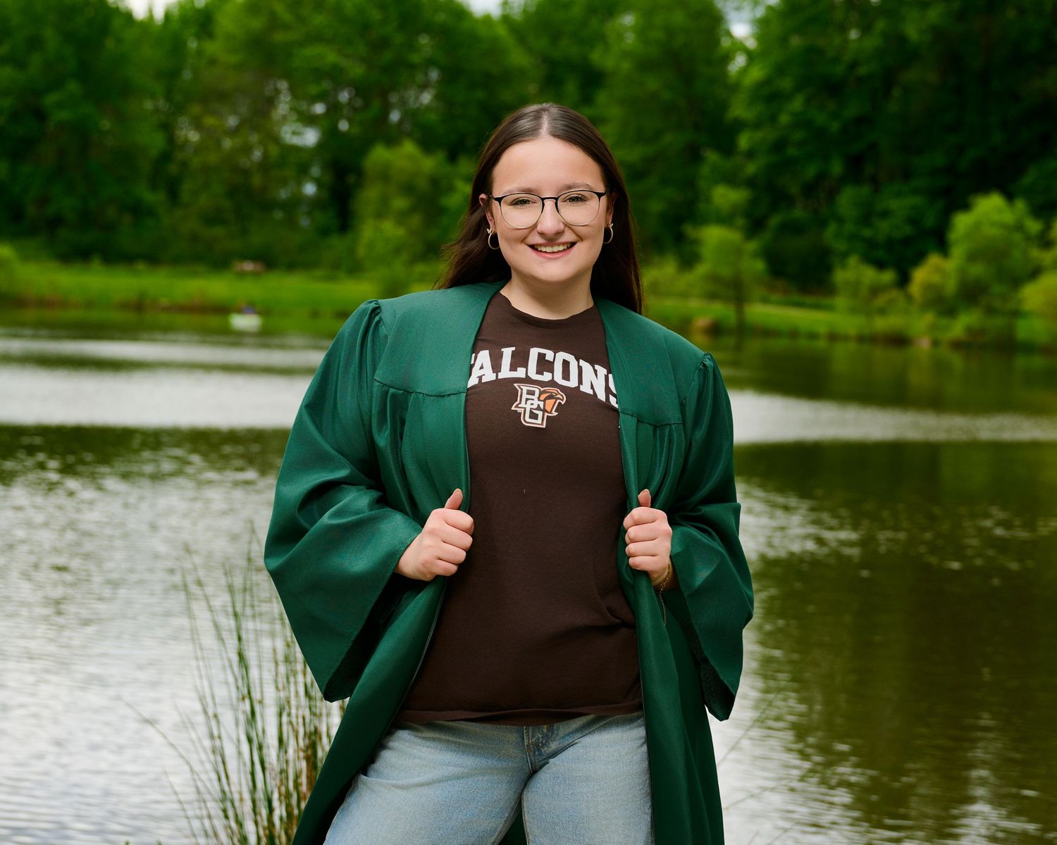 A casual outdoor portrait by a lake featuring someone wearing an Alcorn t-shirt and green graduation gown.