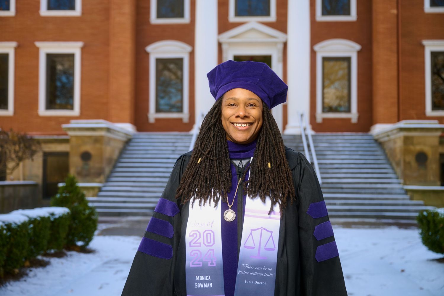 Graduate in doctoral regalia smiles in front of red brick university building in winter.