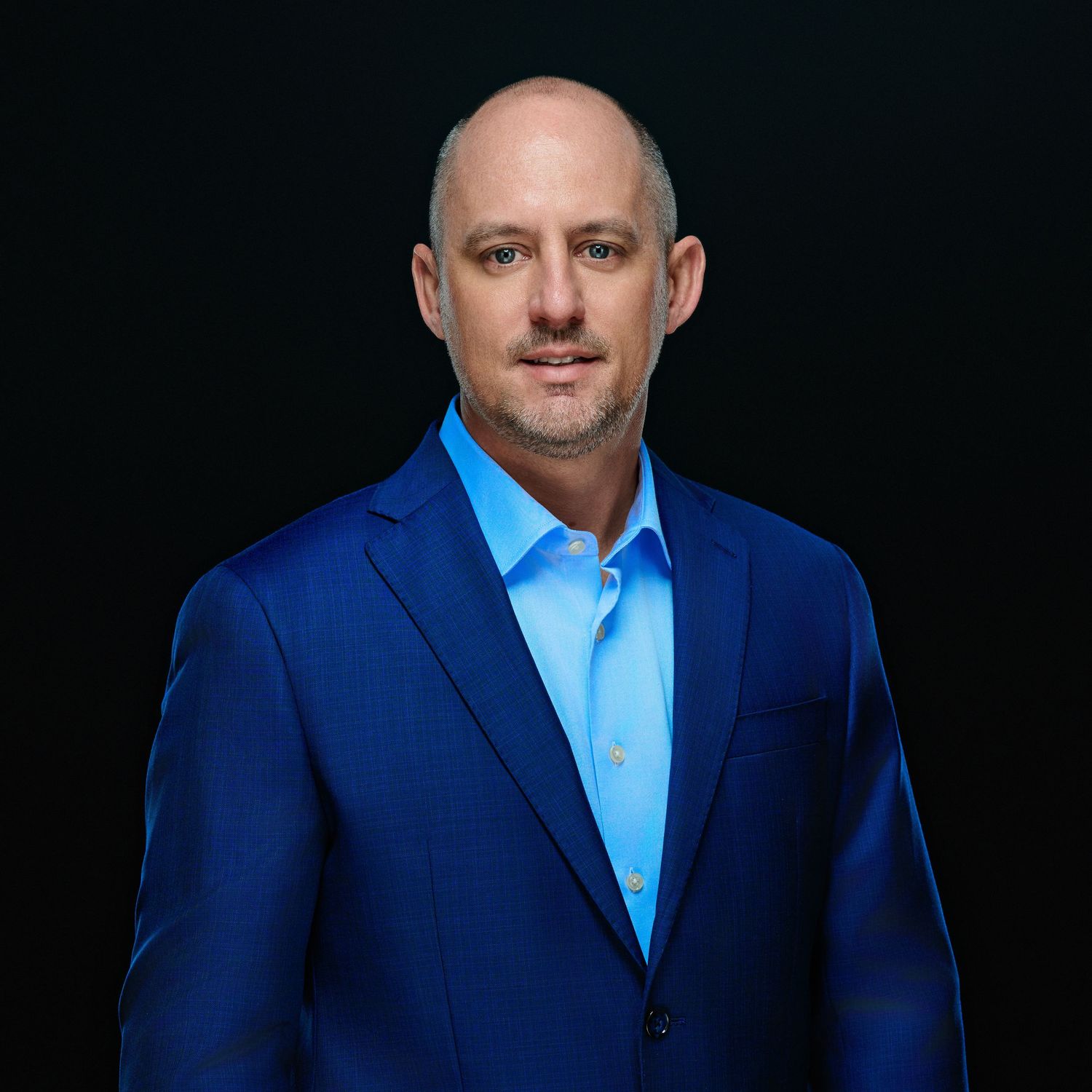 Professional portrait in blue suit jacket and light blue shirt against dark background.