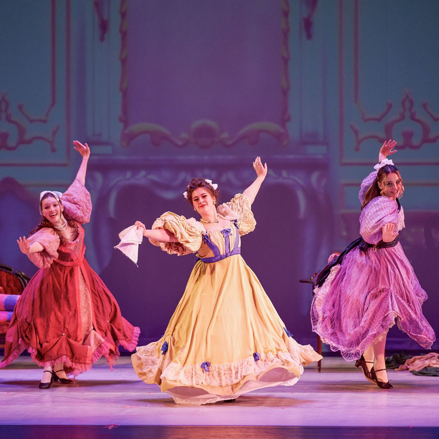 Dancers in colorful period dresses perform a classical dance number on stage.