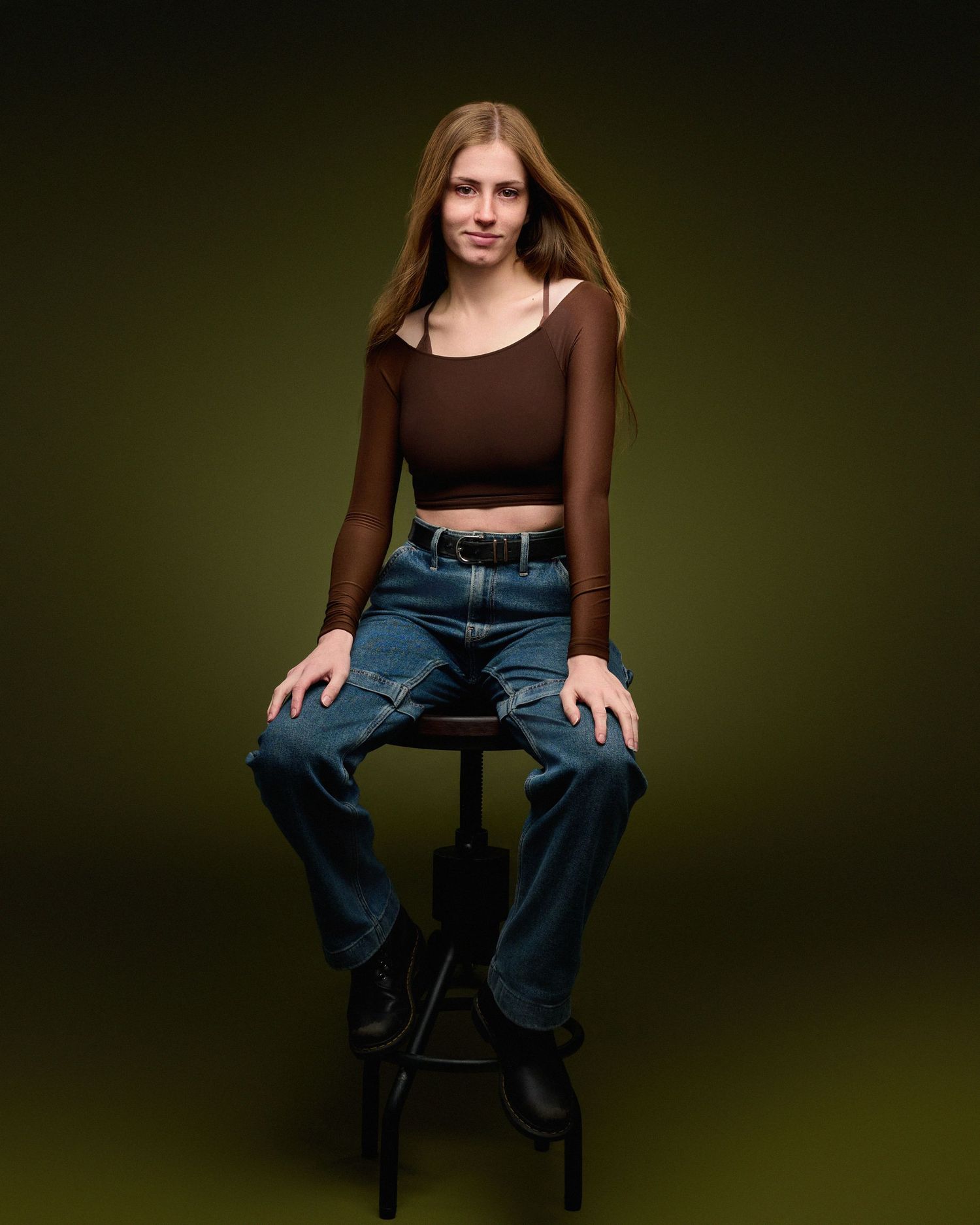 Fashion portrait of a model wearing a brown crop top and jeans while sitting on a black stool against dark backdrop.