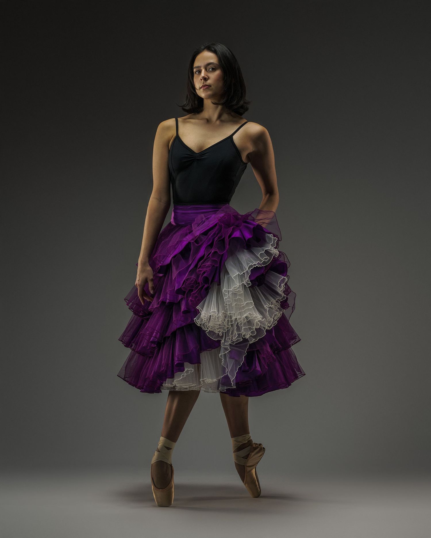 Dancer in black leotard and purple ruffled skirt poses en pointe against dark backdrop.