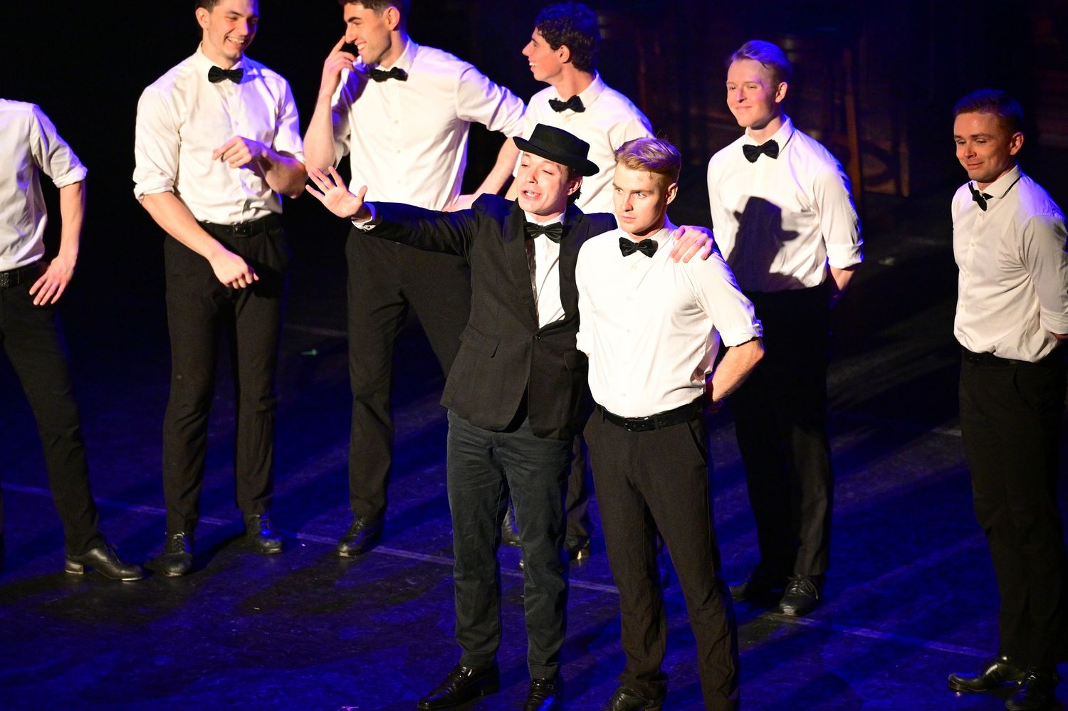 Group of performers in formal black and white attire pose during stage performance.