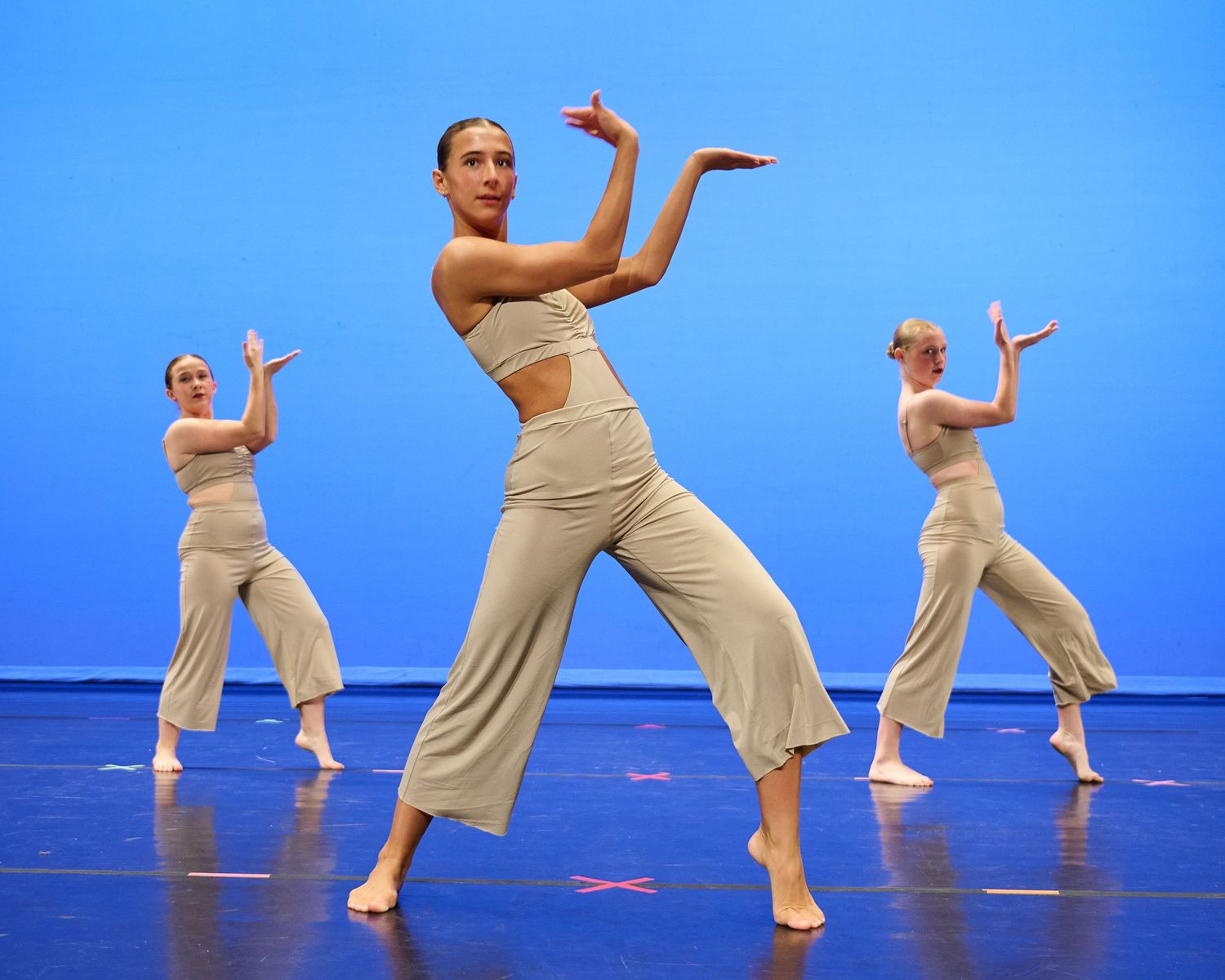 Three dancers in flowing beige pants perform a synchronized modern dance routine against a blue backdrop.