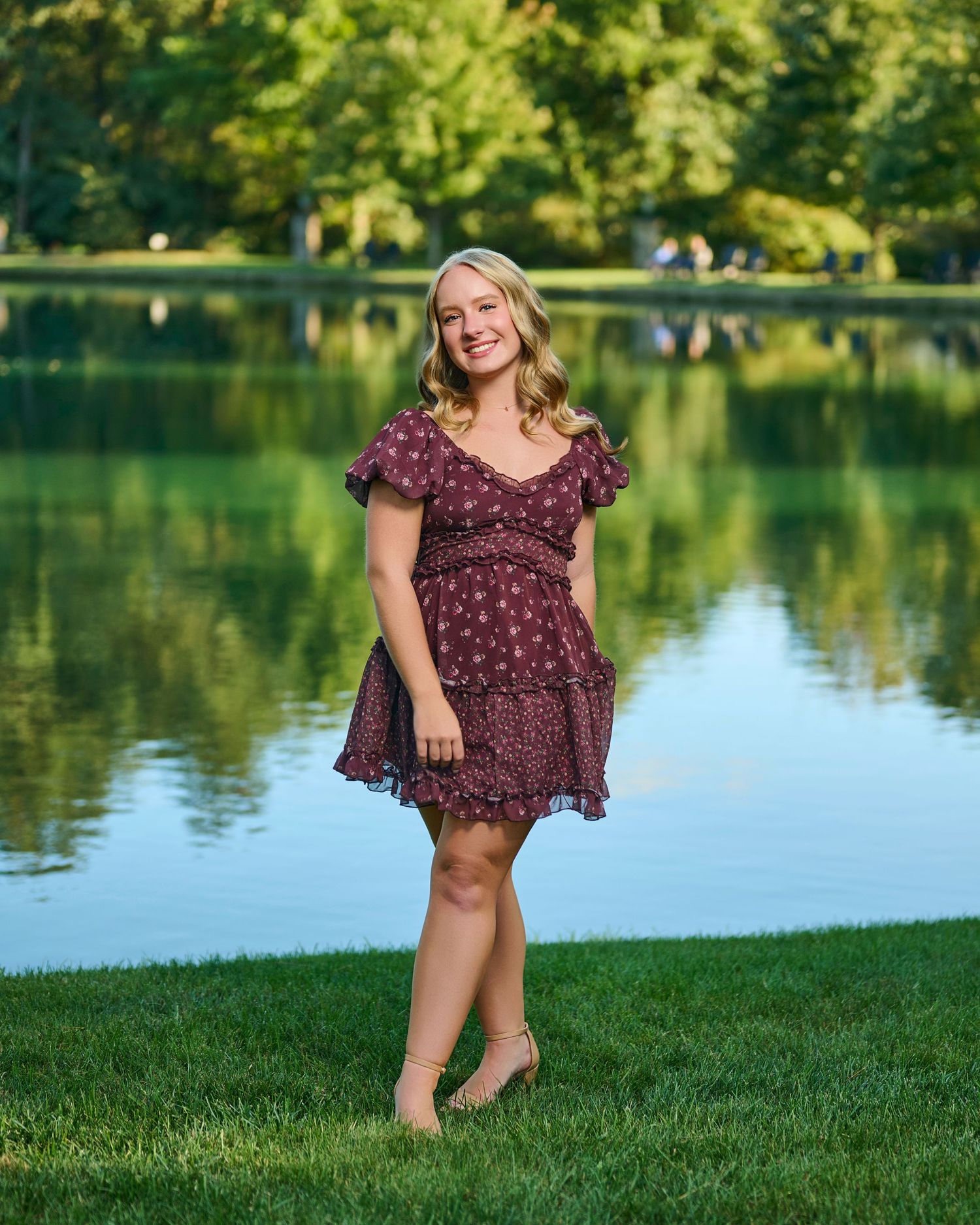 Person in floral burgundy dress poses by serene lake on grassy shore.
