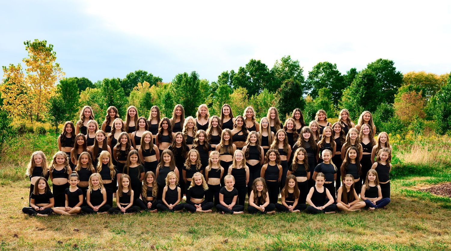 Large group of dancers posed outdoors in matching outfits against natural background.